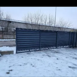 Przesuwna brama przemysłowa z poziomych paneli w kolorze grafitowym, częściowo otwarta, na tle zimowego krajobrazu z budynkiem przemysłowym i drzewami bez liści.