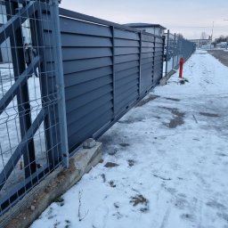 MAZO Creator - Ciemnoszara brama przesuwna z poziomych paneli, widoczna zza metalowej siatki ogrodzeniowej w zimowej scenerii z odśnieżonym chodnikiem i hydrantem.