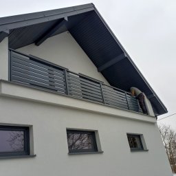 MAZO Creator - Montażysta mocuje poziome panele ogrodzeniowe na balkonie nowoczesnego domu z szarą dachówką, widoczne okna z ciemnymi ramami i fragmenty drzew bez liści.