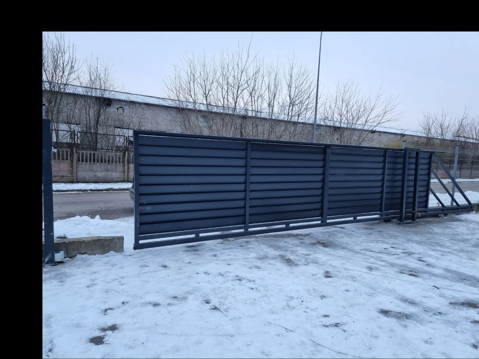 Przesuwna brama przemysłowa z poziomych paneli w kolorze grafitowym, częściowo otwarta, na tle zimowego krajobrazu z budynkiem przemysłowym i drzewami bez liści.