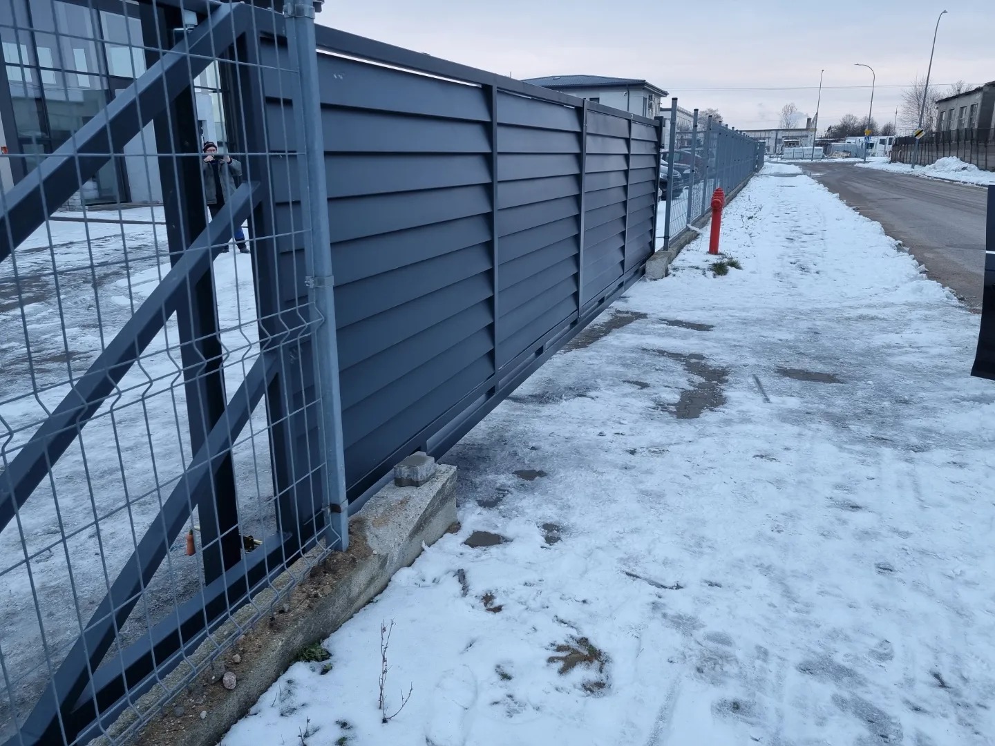 Ciemnoszara brama przesuwna z poziomych paneli, widoczna zza metalowej siatki ogrodzeniowej w zimowej scenerii z odśnieżonym chodnikiem i hydrantem.