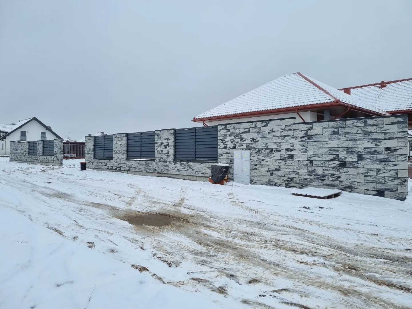 Nowoczesne ogrodzenie z szarego kamienia łupanego i poziomych, grafitowych paneli, widoczne na tle zasypanego śniegiem terenu przed domem.