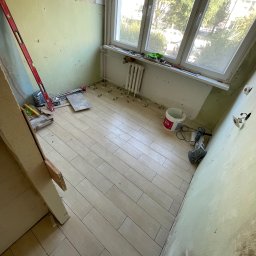 Wojciech Majewski Usługi Remontowo-Budowlane - Ułożone płytki podłogowe imitujące drewno w trakcie remontu balkonu, widoczne narzędzia, poziomica, wiadro z klejem, brakujące listwy przypodłogowe i surowe ściany.