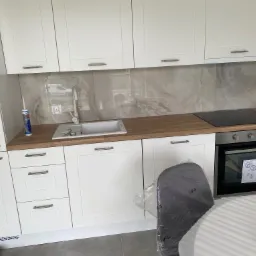 Nowoczesna, jasna kuchnia z białymi szafkami, drewnianym blatem, płytą indukcyjną i piekarnikiem, widoczna tuba z silikonem przy zlewie, na podłodze szare płytki.