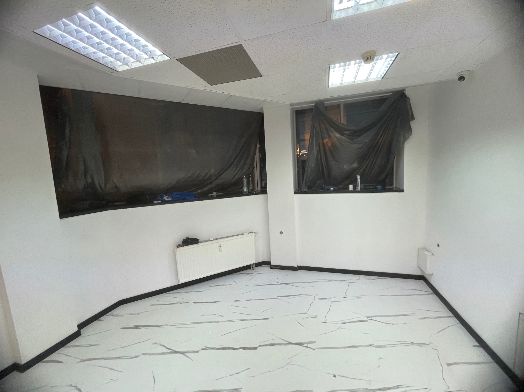 Pusta, biała przestrzeń z oknami zabezpieczonymi czarną folią malarską, grzejnikiem i podłogą w płytkach imitujących marmur, przygotowana do prac wykończeniowych.