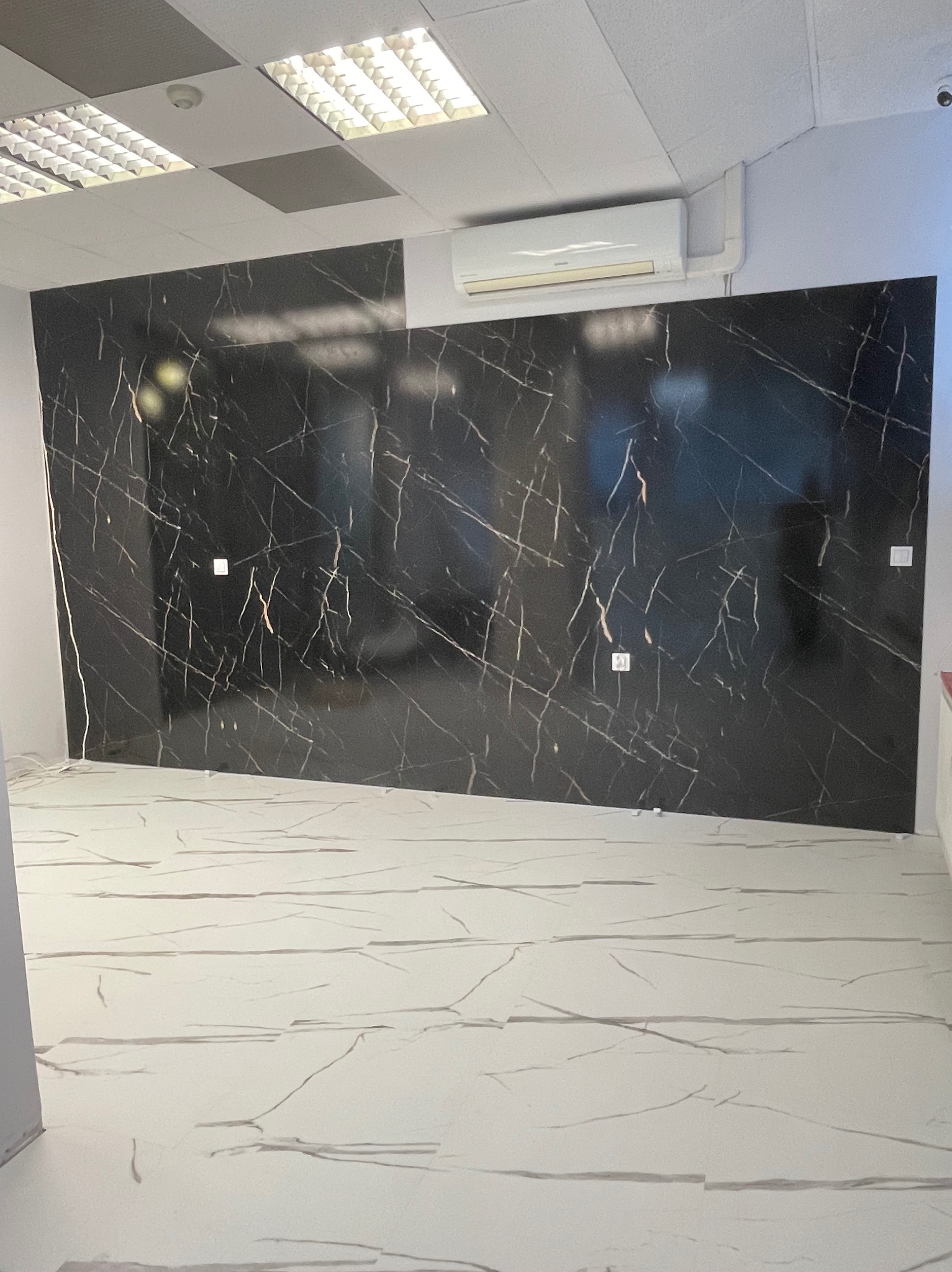 Wykończone wnętrze z czarną ścianą imitującą marmur z białymi żyłami i jasną podłogą z podobnym wzorem, widoczny klimatyzator na ścianie.
