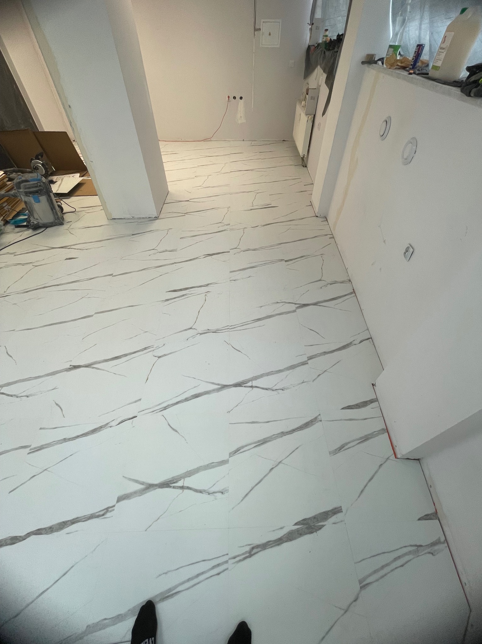 Wnętrze w trakcie wykańczania: białe płytki podłogowe imitujące marmur, kolumna nośna, przygotowane ściany z widocznymi puszkami elektrycznymi i narzędzia w tle.