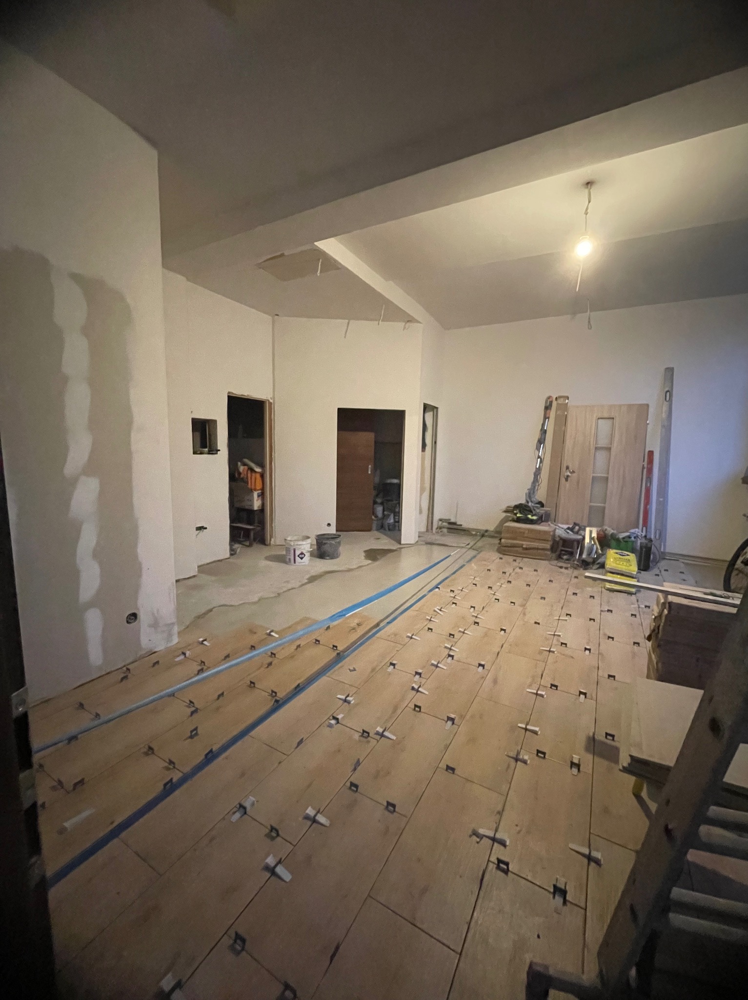 Wnętrze w trakcie remontu: przygotowane podłoże pod parkiet z listwami wyrównującymi i krzyżykami dystansowymi, ściany częściowo wyszpachlowane, widoczne drzwi i narzędzia.