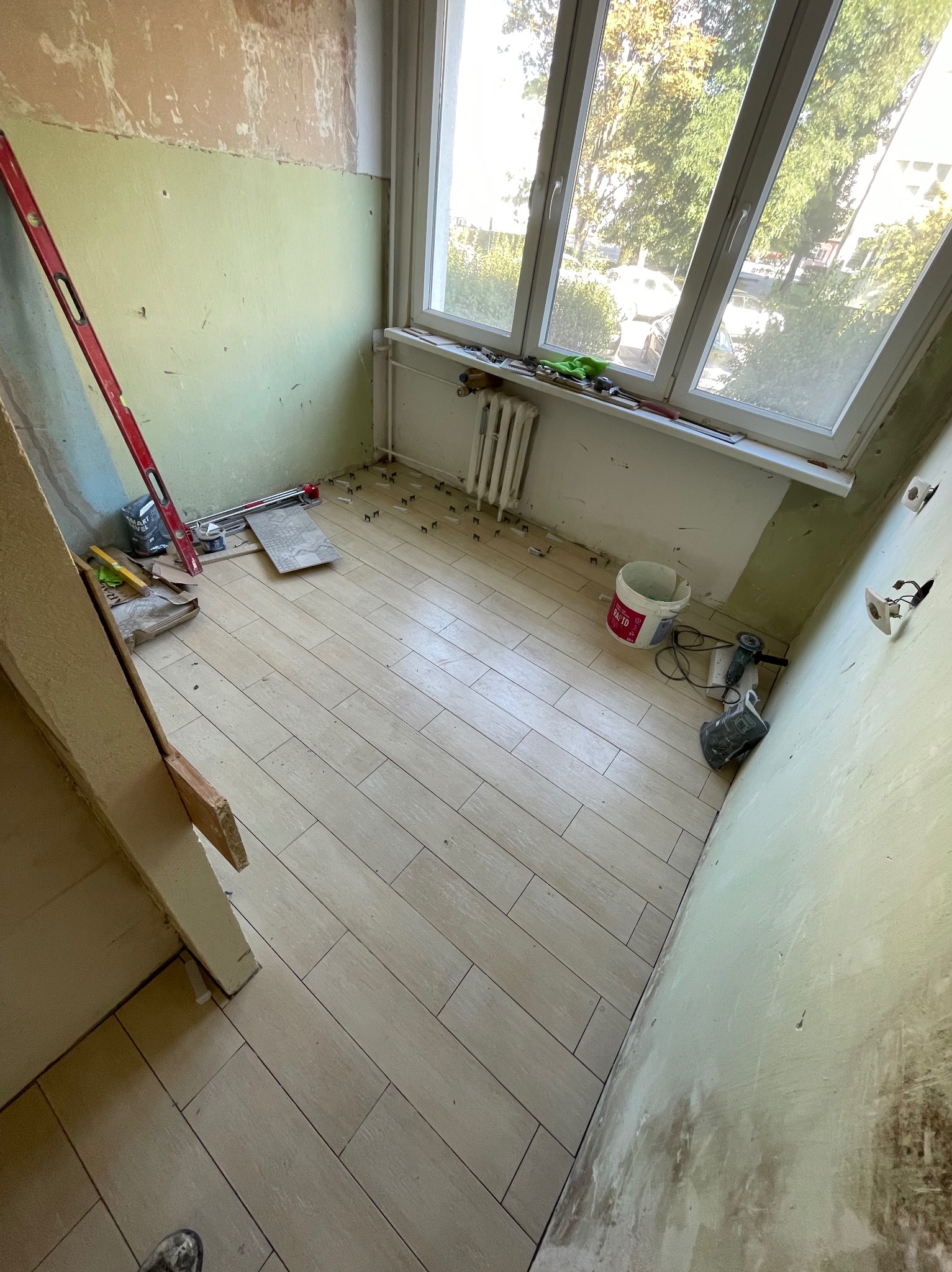 Ułożone płytki podłogowe imitujące drewno w trakcie remontu balkonu, widoczne narzędzia, poziomica, wiadro z klejem, brakujące listwy przypodłogowe i surowe ściany.