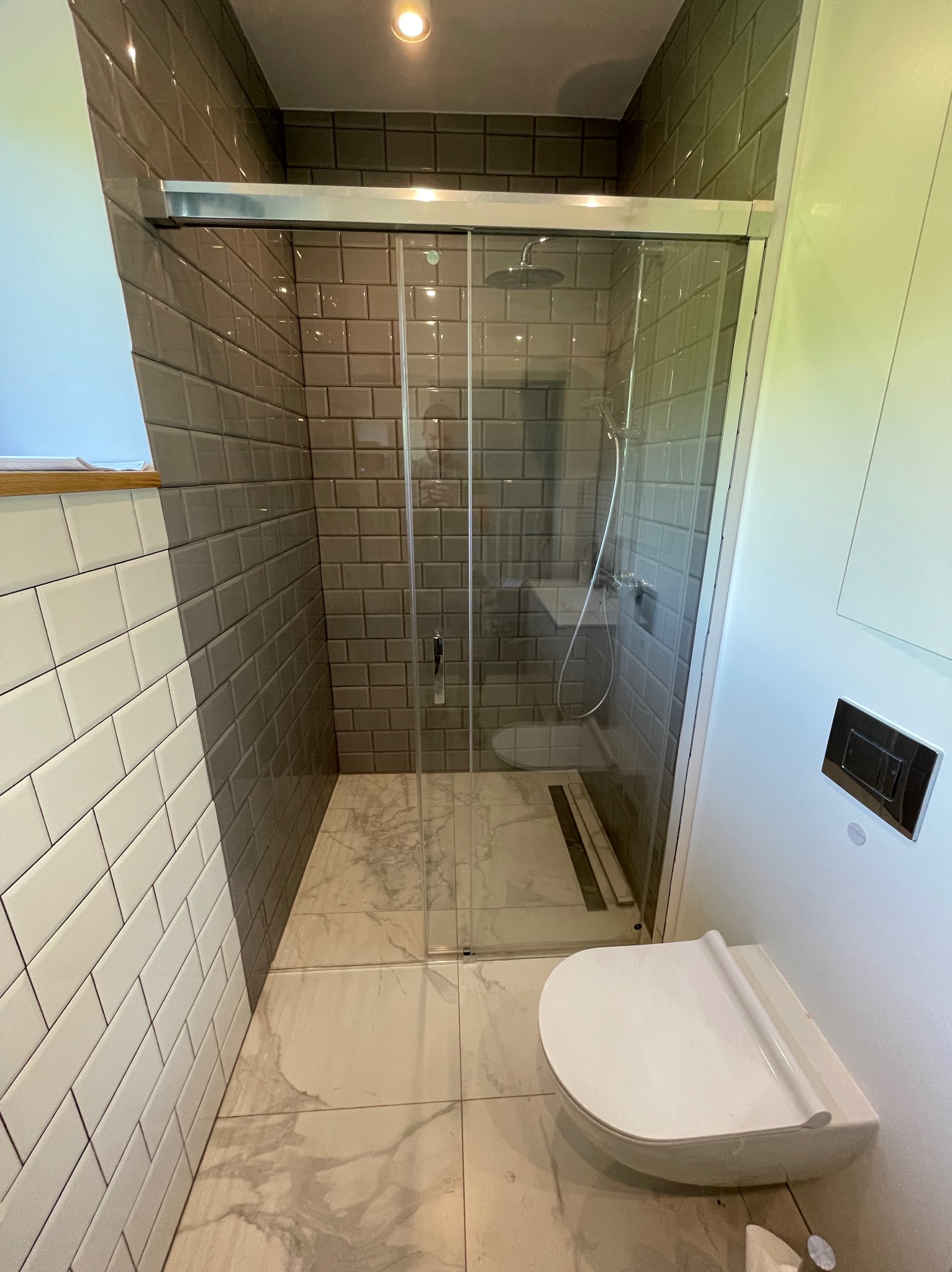 Nowoczesna łazienka z kabiną prysznicową typu walk-in, wyłożona białymi i szarymi płytkami w stylu metro, z białą podwieszaną toaletą i marmurową podłogą.