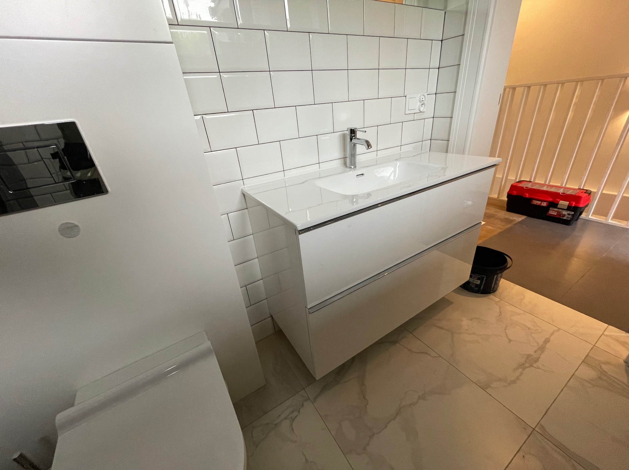 Nowoczesna łazienka z białą umywalką nablatową, chromowaną baterią i białymi płytkami typu metro na ścianie. W tle widoczny czerwony kufer narzędziowy na korytarzu.