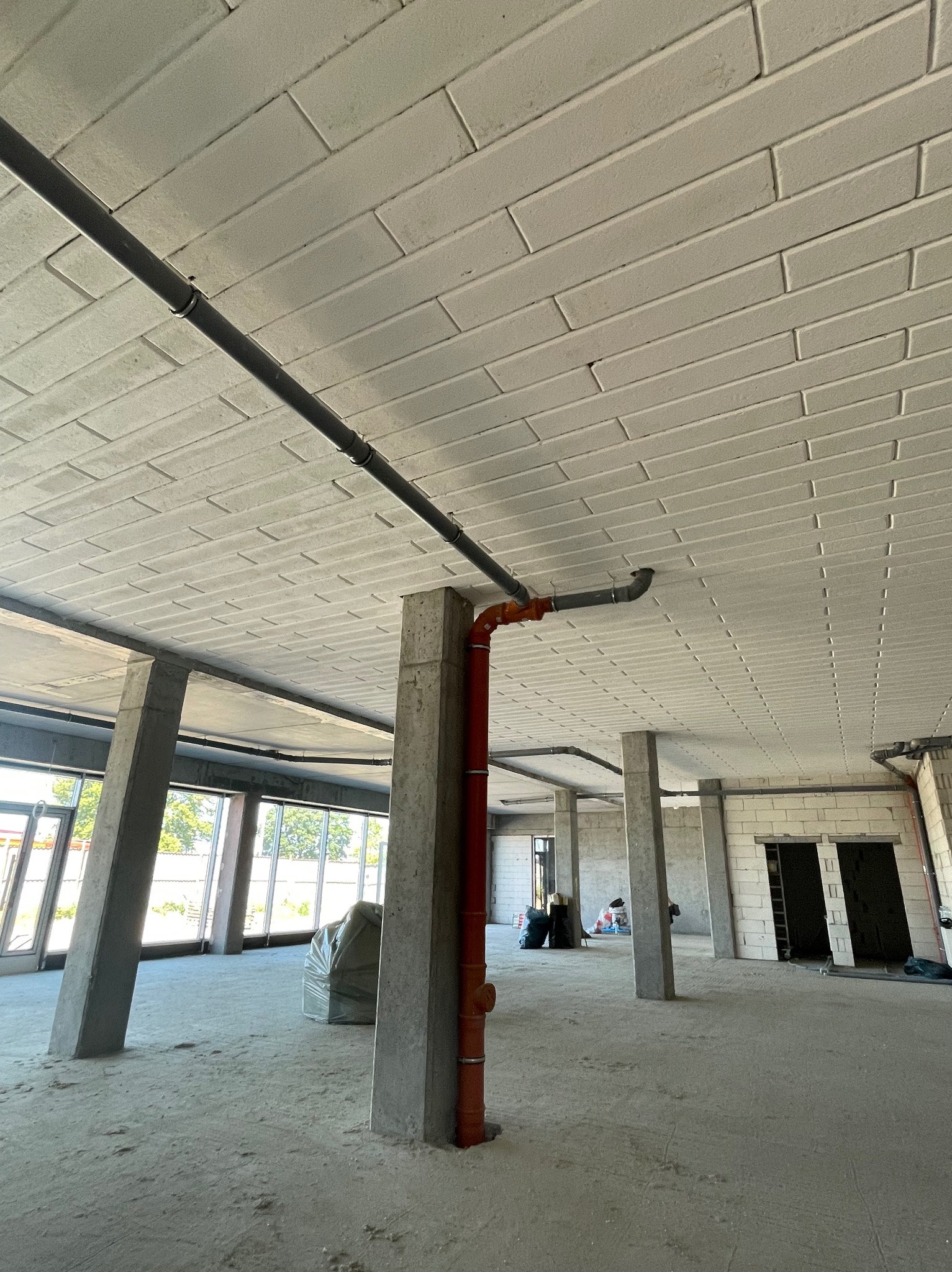 Surowy stan wnętrza budynku z widocznymi betonowymi kolumnami, instalacją rur pod sufitem z bloków, dużymi oknami i workiem materiałów budowlanych.