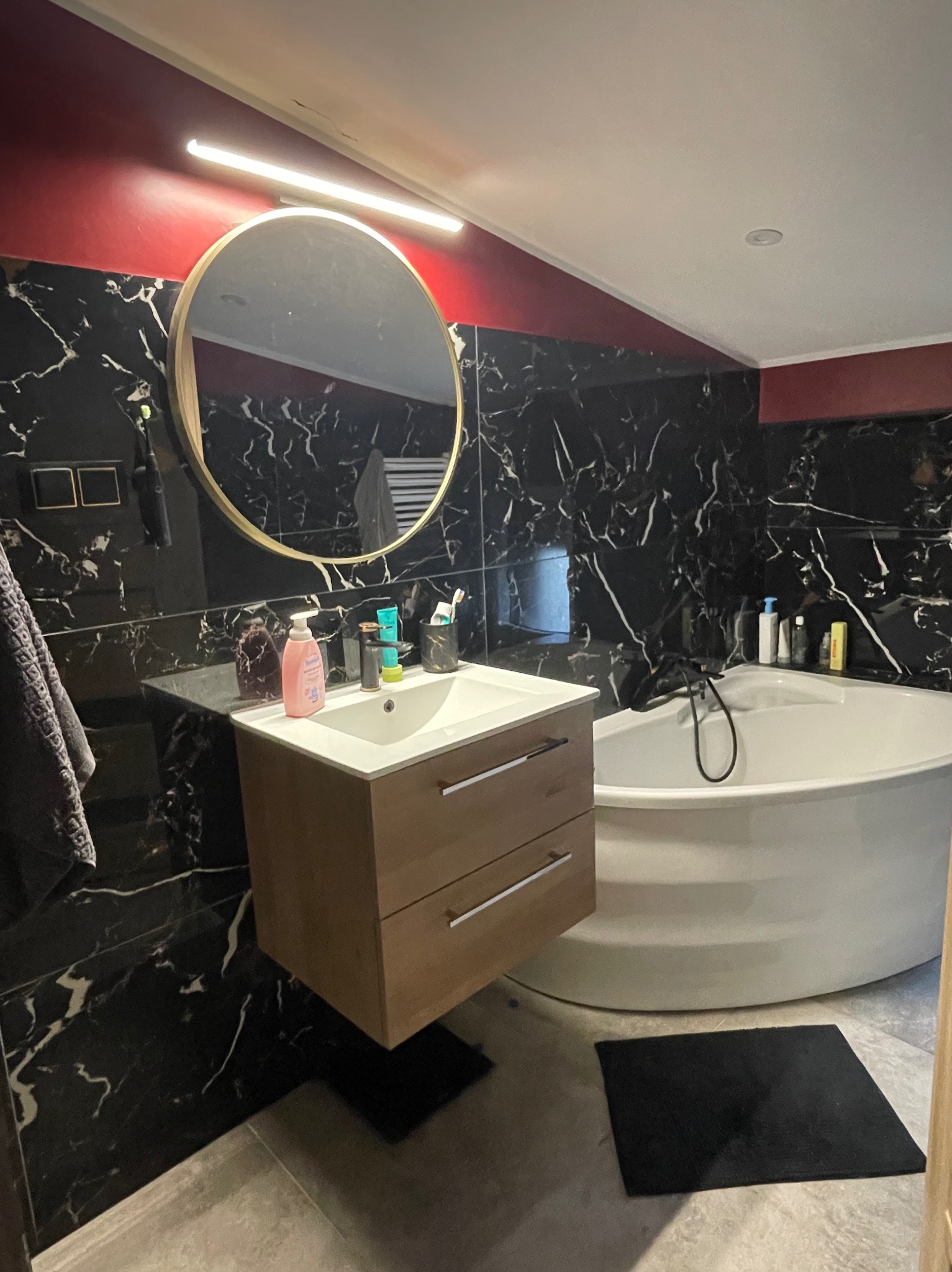 Nowoczesna łazienka z czarnymi marmurowymi ścianami, okrągłym lustrem z oświetleniem LED, wiszącą szafką pod umywalką, białą wanną wolnostojącą i szarymi płytkami podłogowymi.