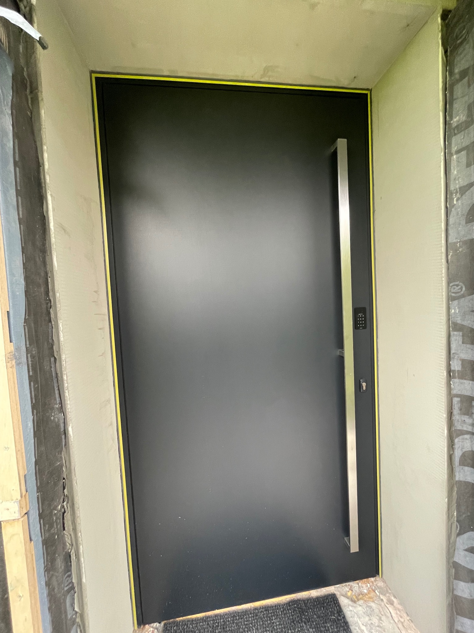 Czarna, nowoczesna brama wejściowa z długim, pionowym uchwytem ze stali nierdzewnej i panelem numerycznym, otoczona żółtą taśmą zabezpieczającą, w trakcie montażu w surowym otworze ściennym.