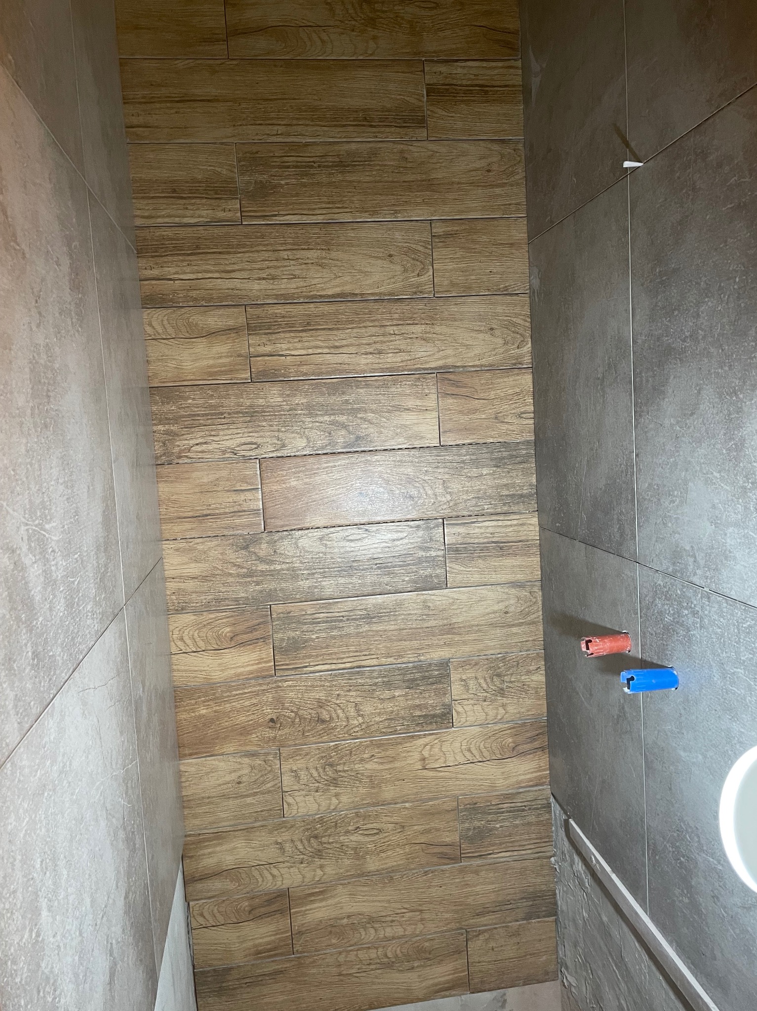 Ściana wyłożona płytkami imitującymi drewno w Grudziądzu, sąsiadująca z szarą ścianą z widocznymi wystającymi rurami z czerwonym i niebieskim zabezpieczeniem.