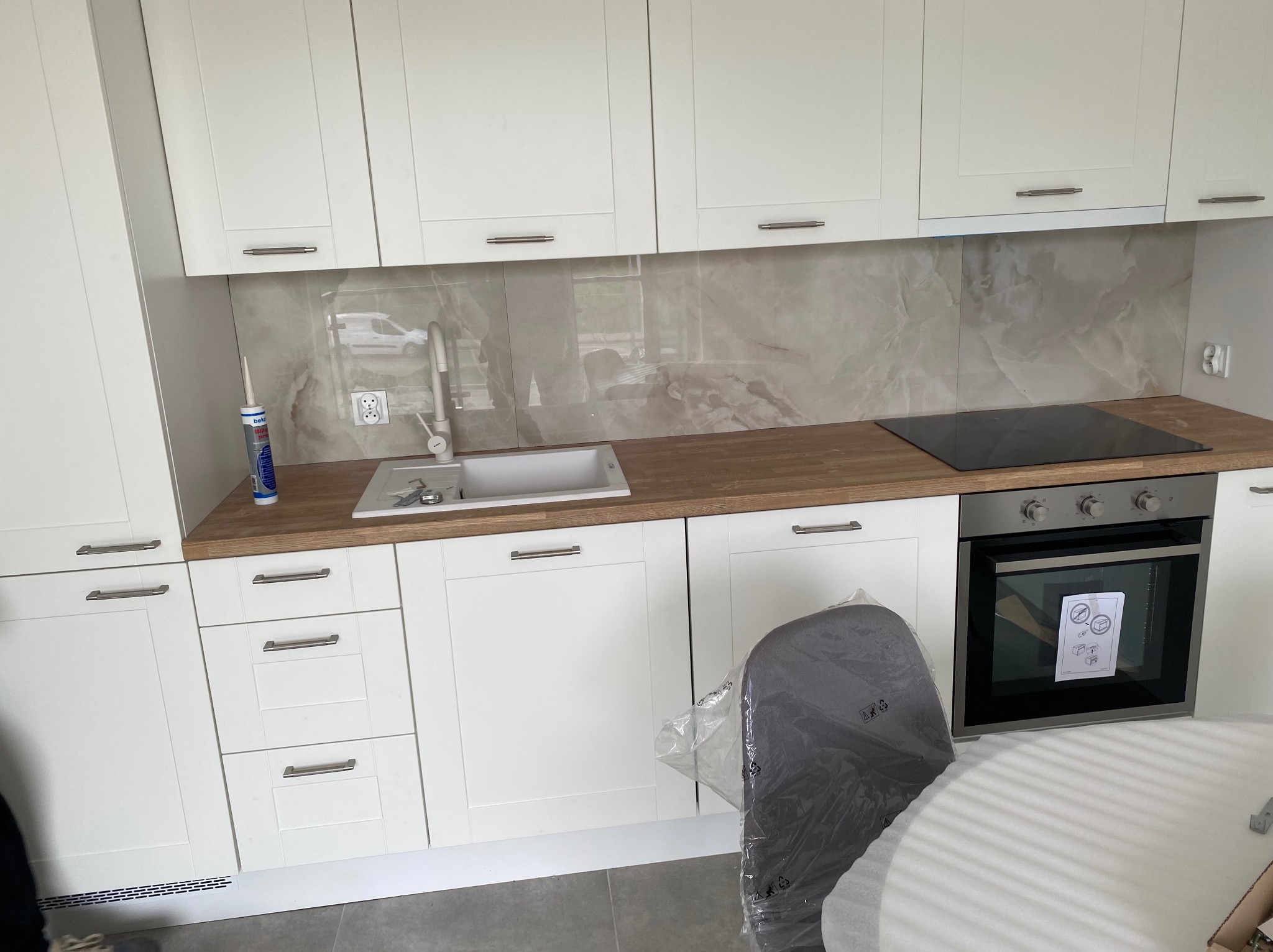 Nowoczesna, jasna kuchnia z białymi szafkami, drewnianym blatem, płytą indukcyjną i piekarnikiem, widoczna tuba z silikonem przy zlewie, na podłodze szare płytki.