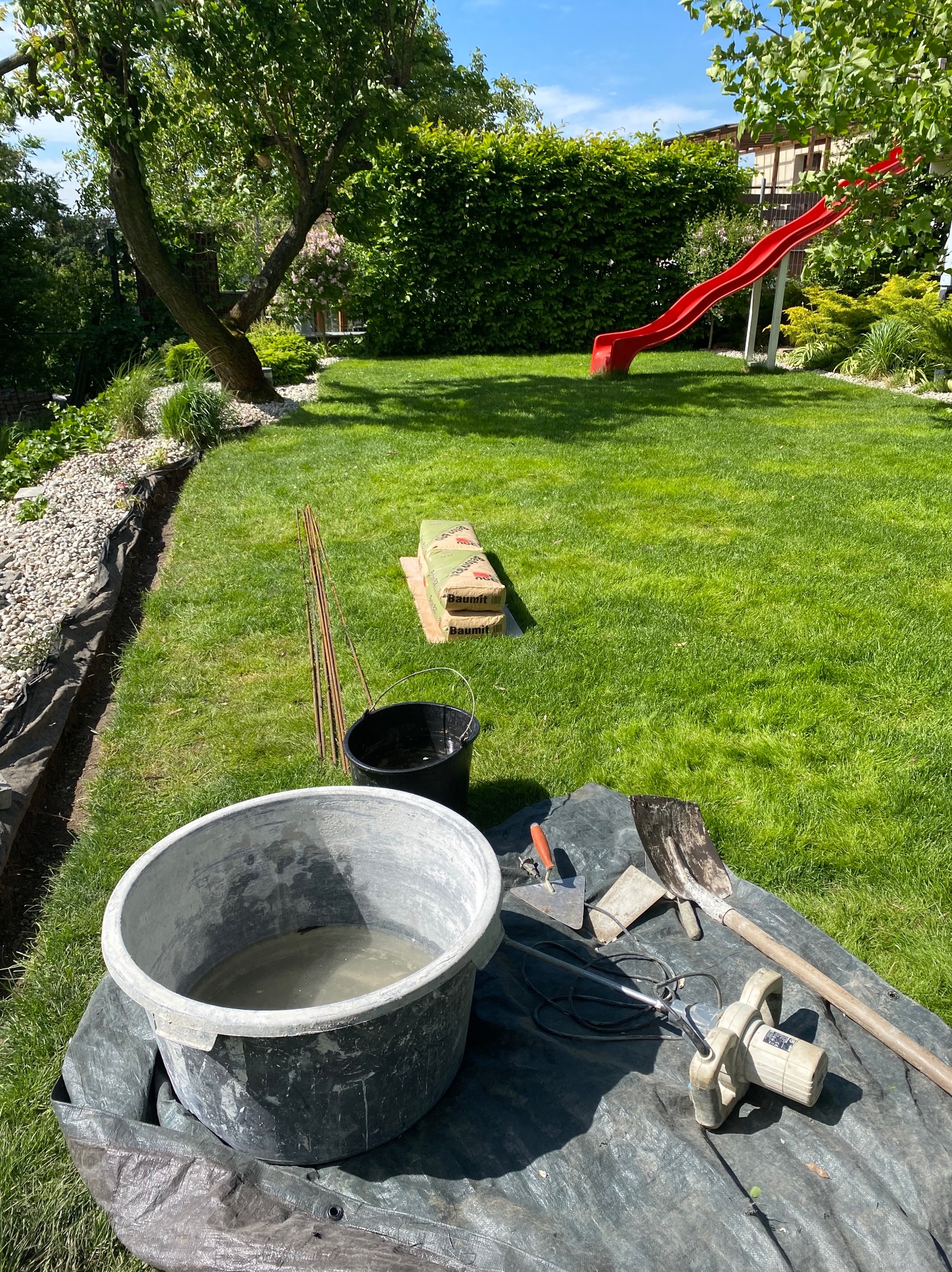 Przygotowanie do prac ziemnych w ogrodzie: wiadro z zaprawą, mieszadło elektryczne, szpadel, kielnia, worki z zaprawą i pręty zbrojeniowe na trawniku, w tle zielony trawnik i czerwona zjeżdżalnia.