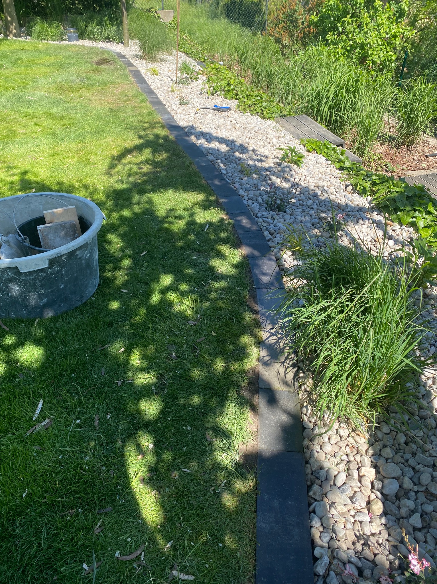 Ułożona obrzeże z ciemnych płyt chodnikowych oddzielająca trawnik od rabaty z białym grysem i roślinnością, widoczny pojemnik budowlany z narzędziami.