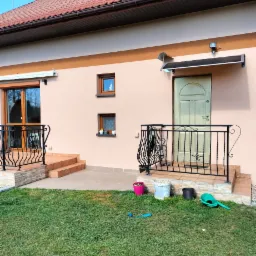 Jasna elewacja domu jednorodzinnego z brązowym dachem, okienkami i wejściem z ozdobną, czarną balustradą, widoczne doniczki i zabawki na trawniku.