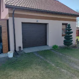 Jasny budynek garażu z brązową bramą segmentową, brązowym pasem dekoracyjnym pod dachem i małym oknem z roletami, na tle zielony trawnik i dekoracyjne drzewko iglaste.