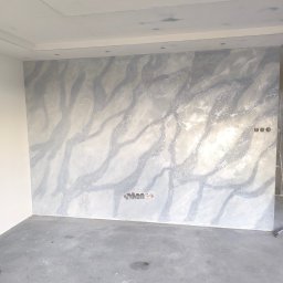 Mar Bud - Ściana z dekoracyjnym tynkiem w odcieniach szarości i bieli, imitującym marmurkowy wzór. Widoczne puszki elektryczne i drabina w rogu. Podłoga betonowa.
