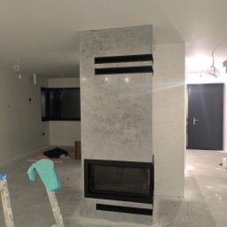 Mar Bud - Wykończony kominek z dekoracyjnym tynkiem w szarościach, czarne elementy ozdobne. W tle widoczne okno i drzwi, pomieszczenie w trakcie wykańczania.