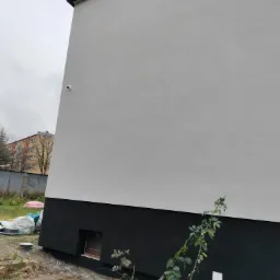 Świeżo otynkowana i pomalowana elewacja budynku z widoczną kamerą monitoringu, czarnym cokołem i młodą rośliną pnącą się wzdłuż ściany.