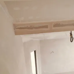 Wnętrze w trakcie prac wykończeniowych: sufit z zamontowanymi profilami i miejscowymi poprawkami gipsowymi, ściany po gruntowaniu, widoczne kable elektryczne, okno z parapetem i fragment drabiny.