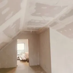 Poddasze w trakcie wykańczania: ściany i sufit pokryte gładzią gipsową, widoczne kable, listwy dekoracyjne pod sufitem, otwarte przejście do kolejnego pomieszczenia z ułożonymi deskami.