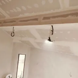 Wnętrze w trakcie prac wykończeniowych; widoczny pracownik z kielnią szpachlową, szpachlowana ściana i sufit z widocznymi łączeniami płyt gipsowo-kartonowych oraz lampa budowlana na statywie.