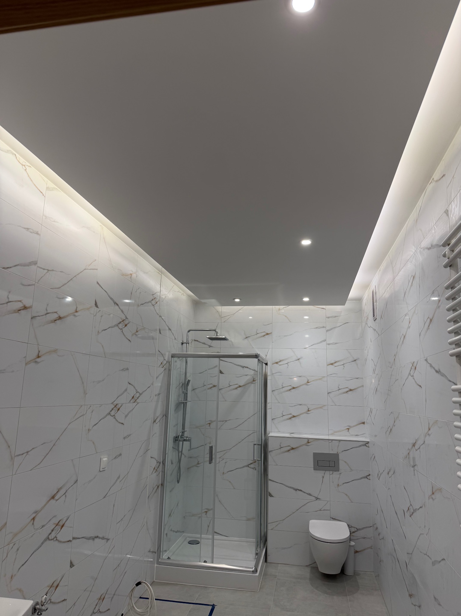 Wykończona łazienka z kabiną prysznicową, toaletą podwieszaną i grzejnikiem drabinkowym. Białe płytki imitujące marmur z brązowymi żyłami. Oświetlenie sufitowe LED.