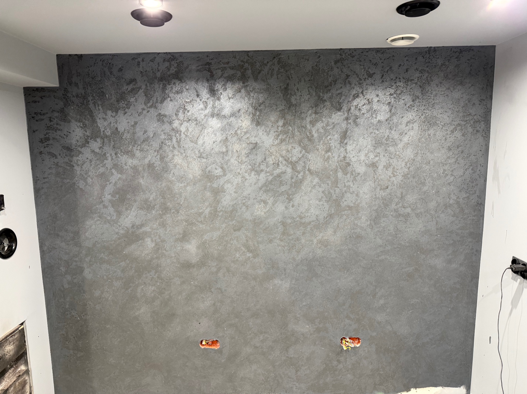 Ściana z dekoracyjnym, szarym tynkiem strukturalnym, widoczne puszki elektryczne. Oświetlenie sufitowe punktowe. Minimalistyczne wykończenie wnętrza.