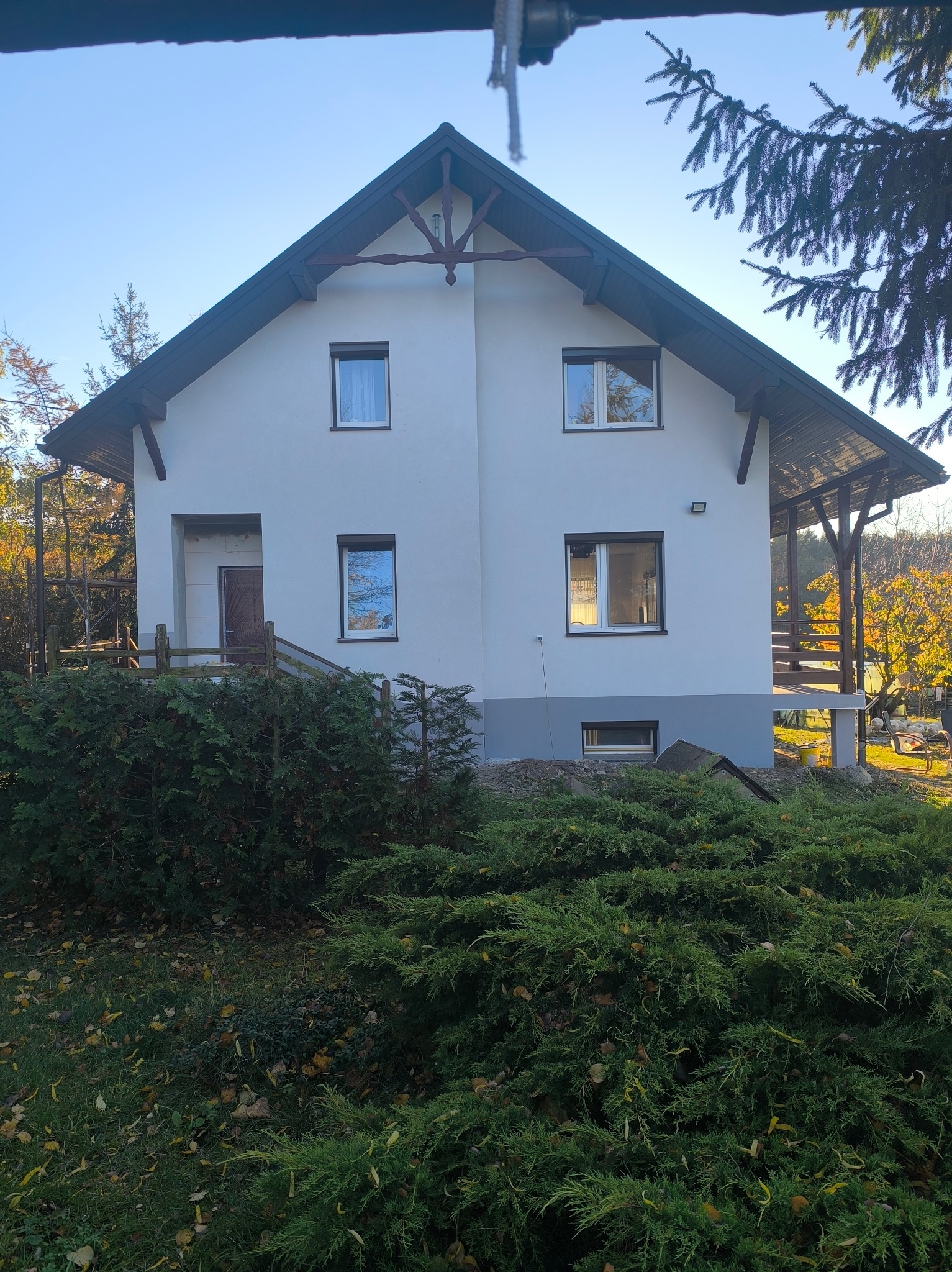 Dwukondygnacyjny dom z białymi ścianami i brązowymi oknami, otoczony zielenią. Widoczny fragment elewacji z drewnianymi elementami i zadaszony taras.