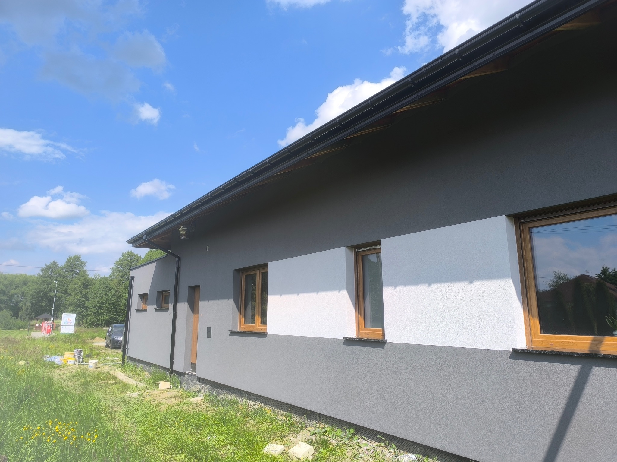 Dwukolorowa elewacja domu jednorodzinnego z drewnianą stolarką okienną, widok z boku, w tle zieleń i błękitne niebo z chmurami. Część elewacji w kolorze szarym, część biała.