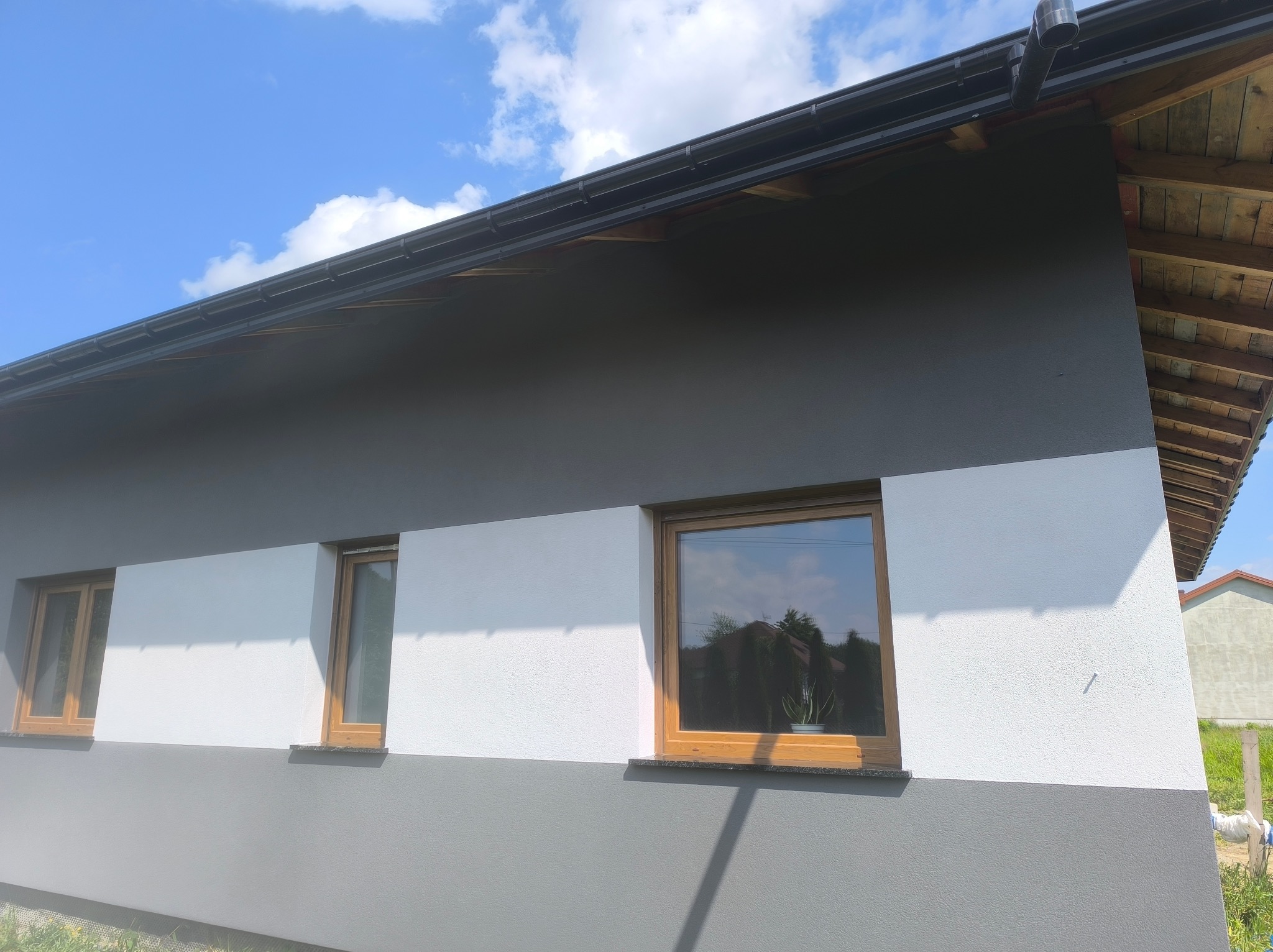 Dwukolorowa elewacja domu z drewnianymi oknami w słoneczny dzień. Szara podstawa i góra, biały pas pośrodku. Widoczne rynny i fragment dachu. W oknach odbicie otoczenia.