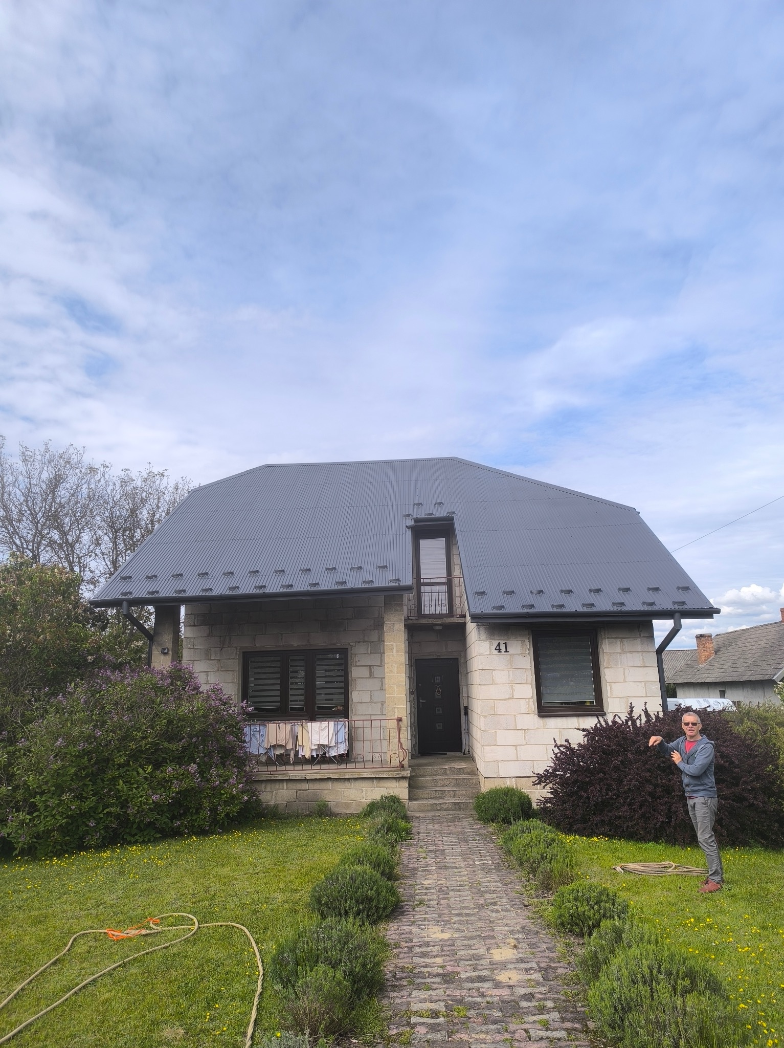 Dom z szarej cegły z nowym, ciemnoszarym dachem z blachy trapezowej. Mężczyzna w okularach słonecznych stoi obok krzewu, wskazując na elewację. Widoczny ogród i błękitne niebo.