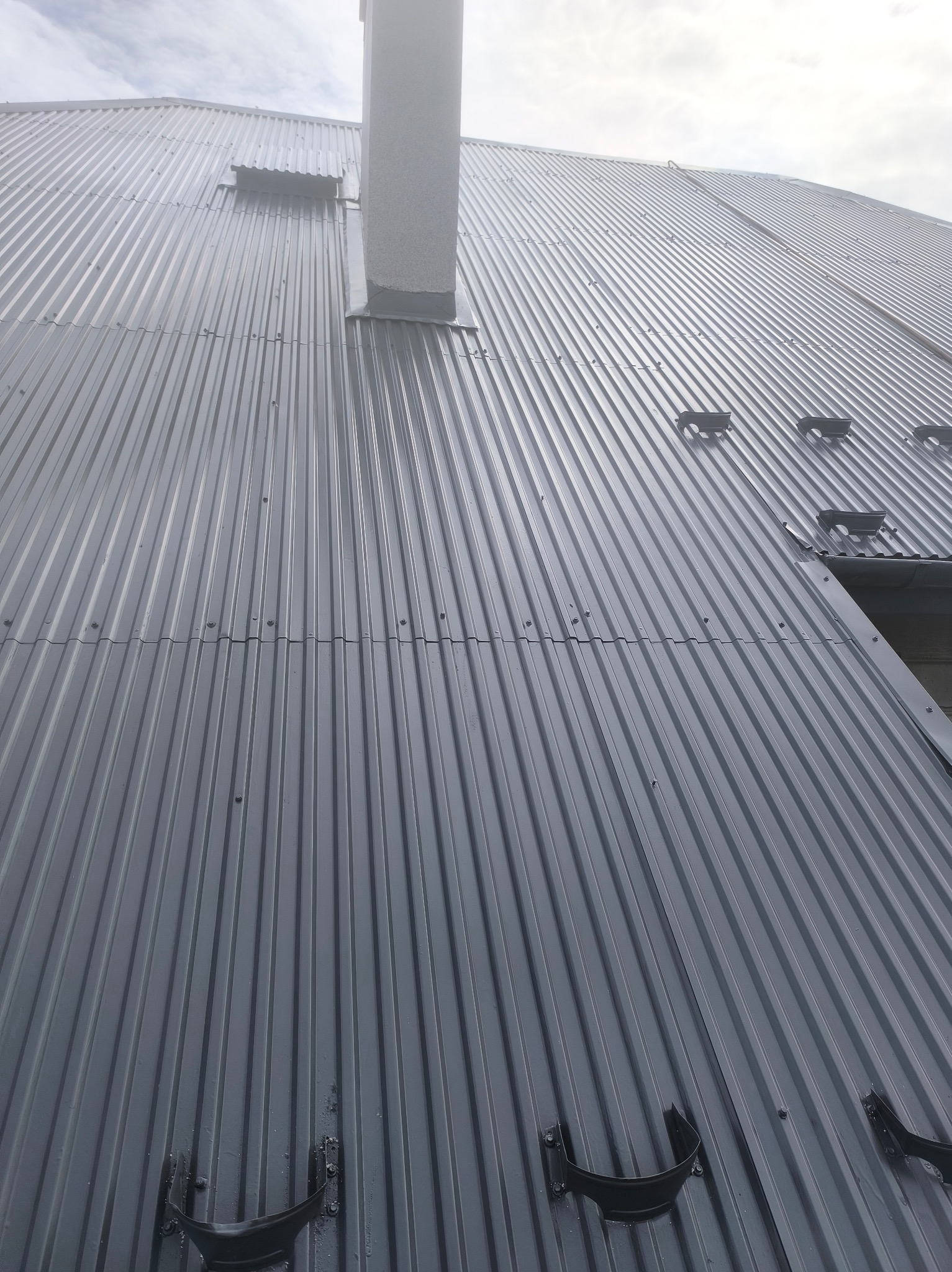 Szary, blaszany dach z kominem i elementami wentylacyjnymi, sfotografowany pod kątem, ukazujący strukturę i detale pokrycia.