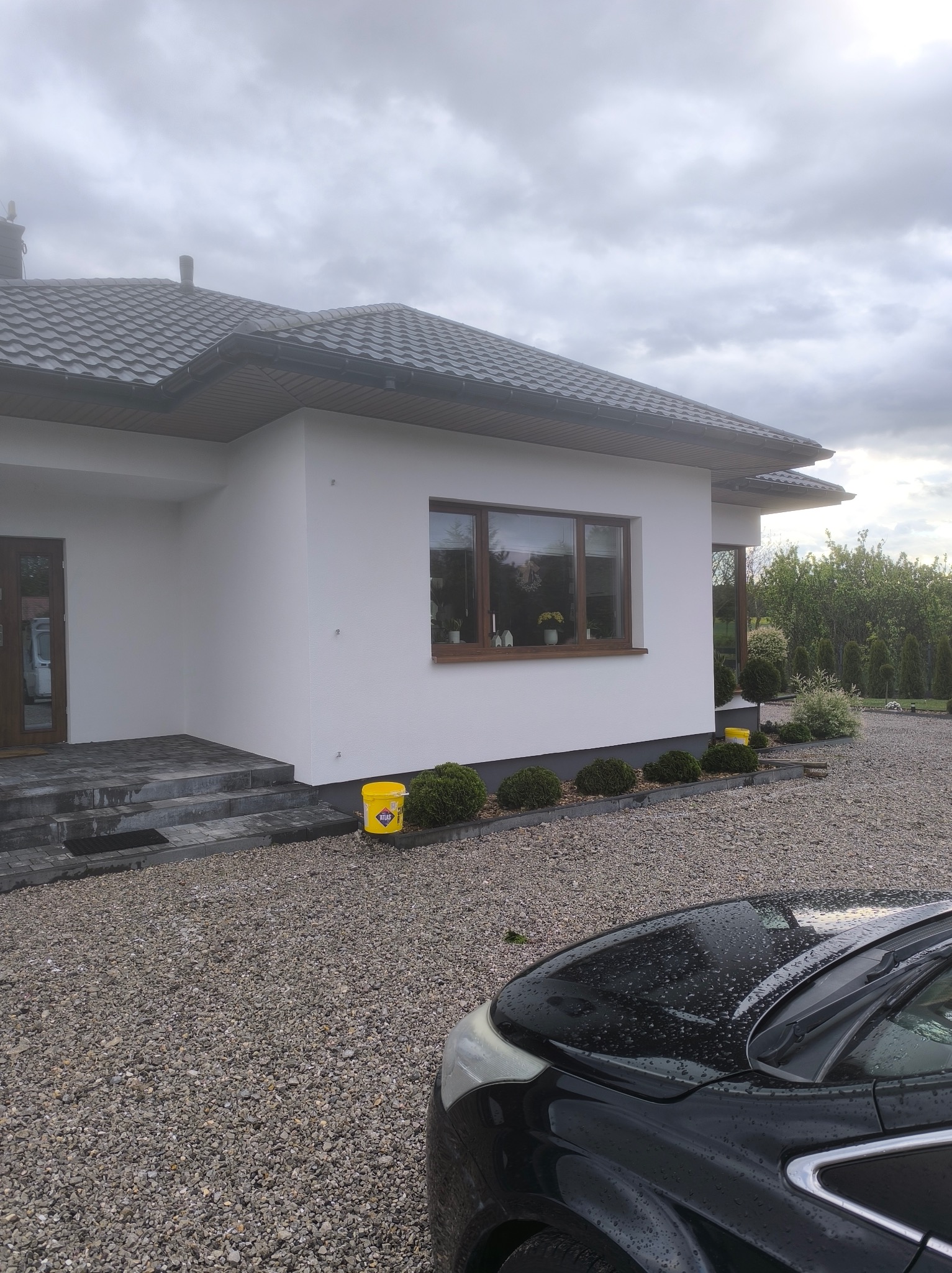 Biały dom jednorodzinny z szarym dachem, widok z boku, z żółtym wiadrem farby Atlas przy ścianie. Część samochodu na pierwszym planie, szara nawierzchnia.
