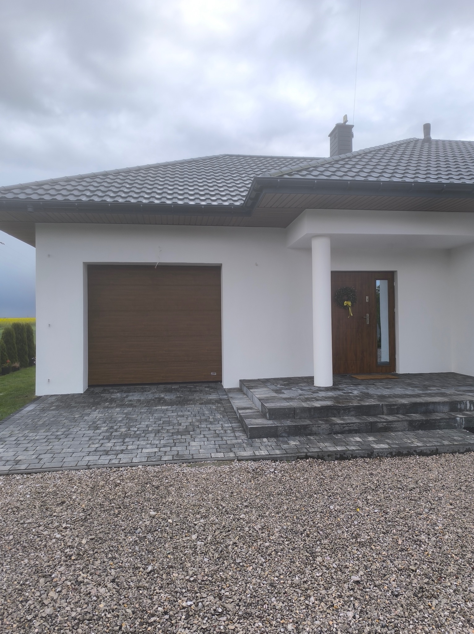 Nowoczesny dom jednorodzinny z brązową bramą garażową i drzwiami wejściowymi. Biała elewacja, szara kostka brukowa i kamień ozdobny. W tle żółte pole.