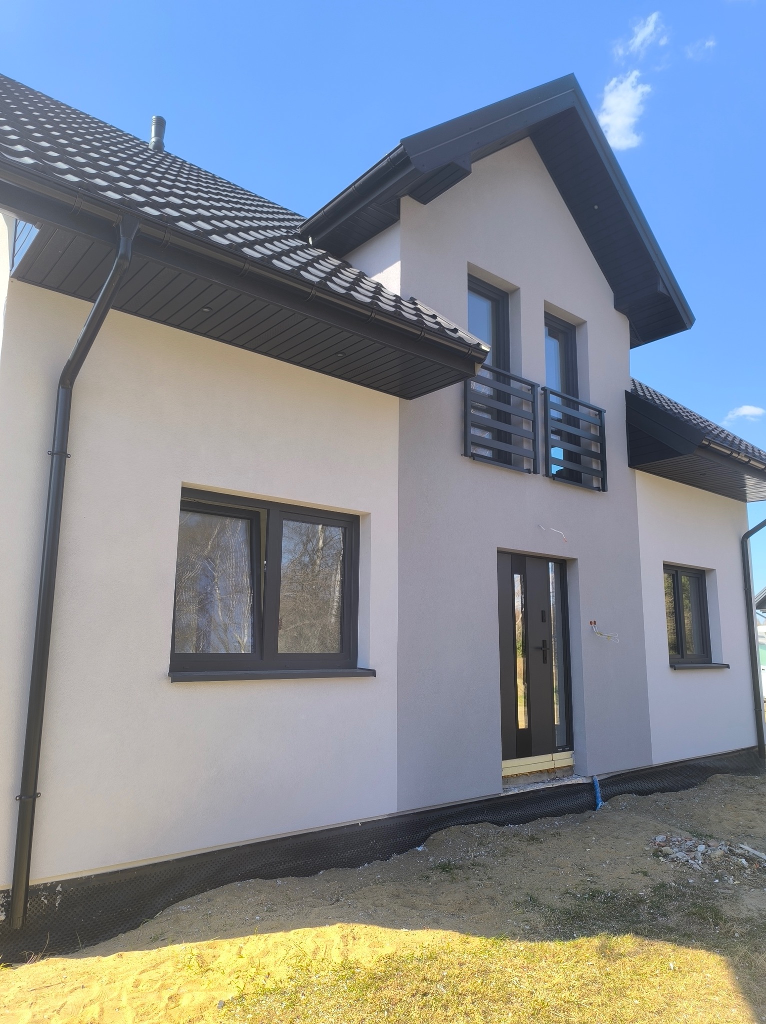 Nowoczesny dom jednorodzinny z jasną elewacją, ciemną stolarką okienną i dachem. Widoczne detale wykończenia, takie jak rynny i balustrada balkonowa.