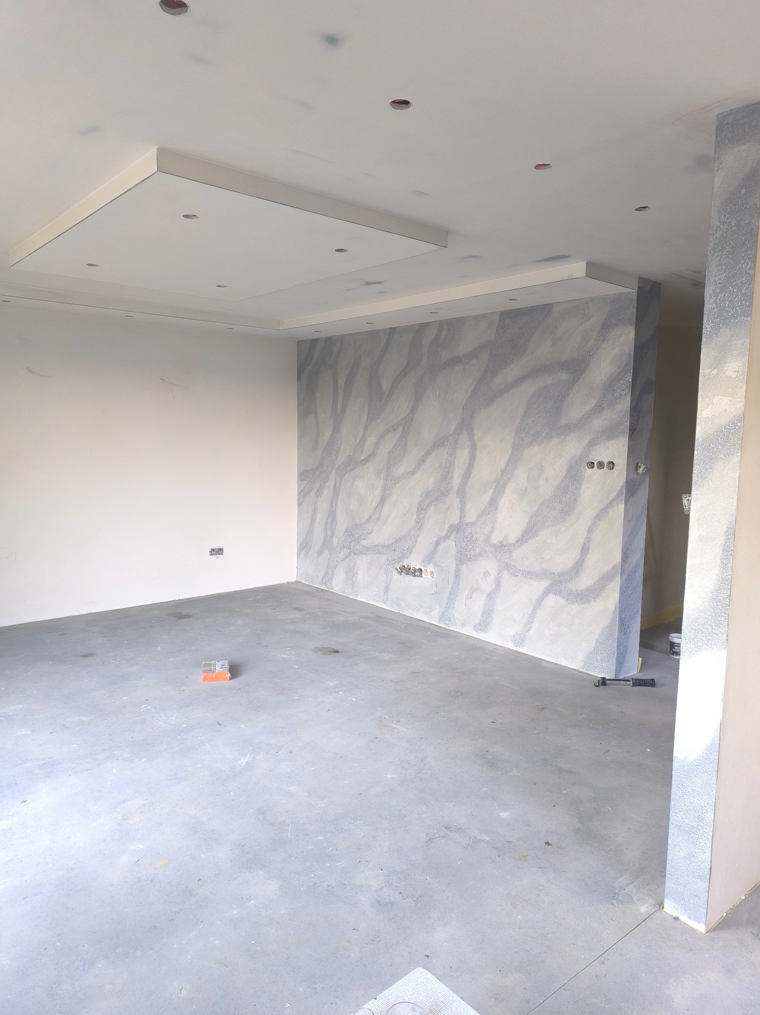 Wnętrze w trakcie wykańczania: ściana z dekoracyjnym wzorem imitującym marmur, surowa podłoga, widoczne puszki elektryczne i narzędzia. Oświetlenie sufitowe w zabudowie.