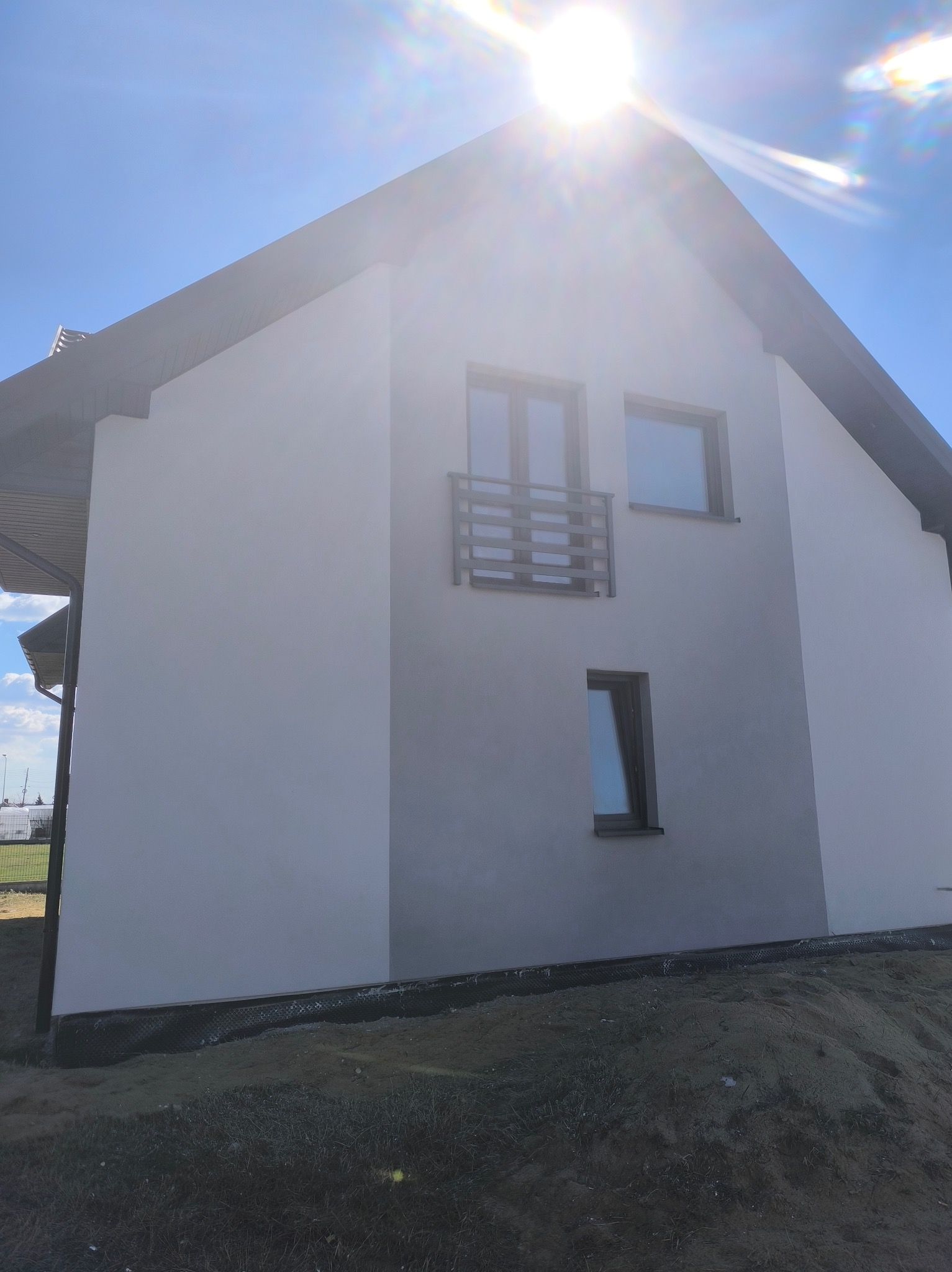 Boczny widok domu z jasną elewacją w słoneczny dzień. Widoczne okna, mały balkon i fragment dachu. Teren wokół domu częściowo przygotowany pod zagospodarowanie.