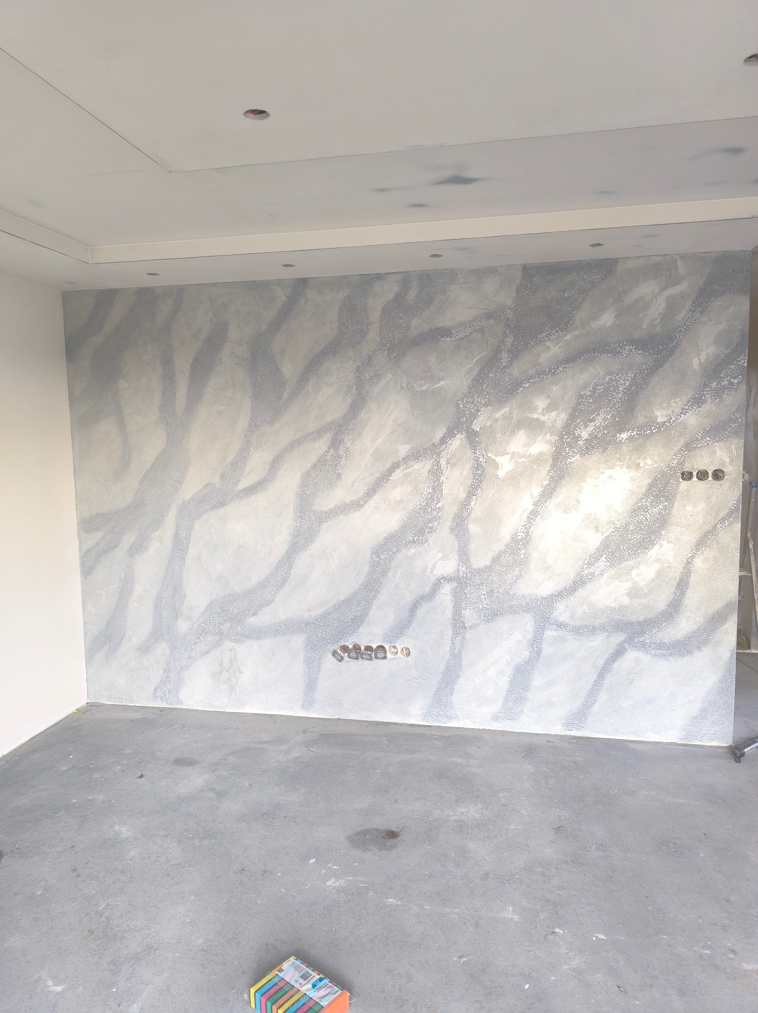 Ściana z dekoracyjnym tynkiem w odcieniach szarości i bieli, imitującym marmurkowy wzór. Widoczne puszki elektryczne i drabina w rogu. Podłoga betonowa.