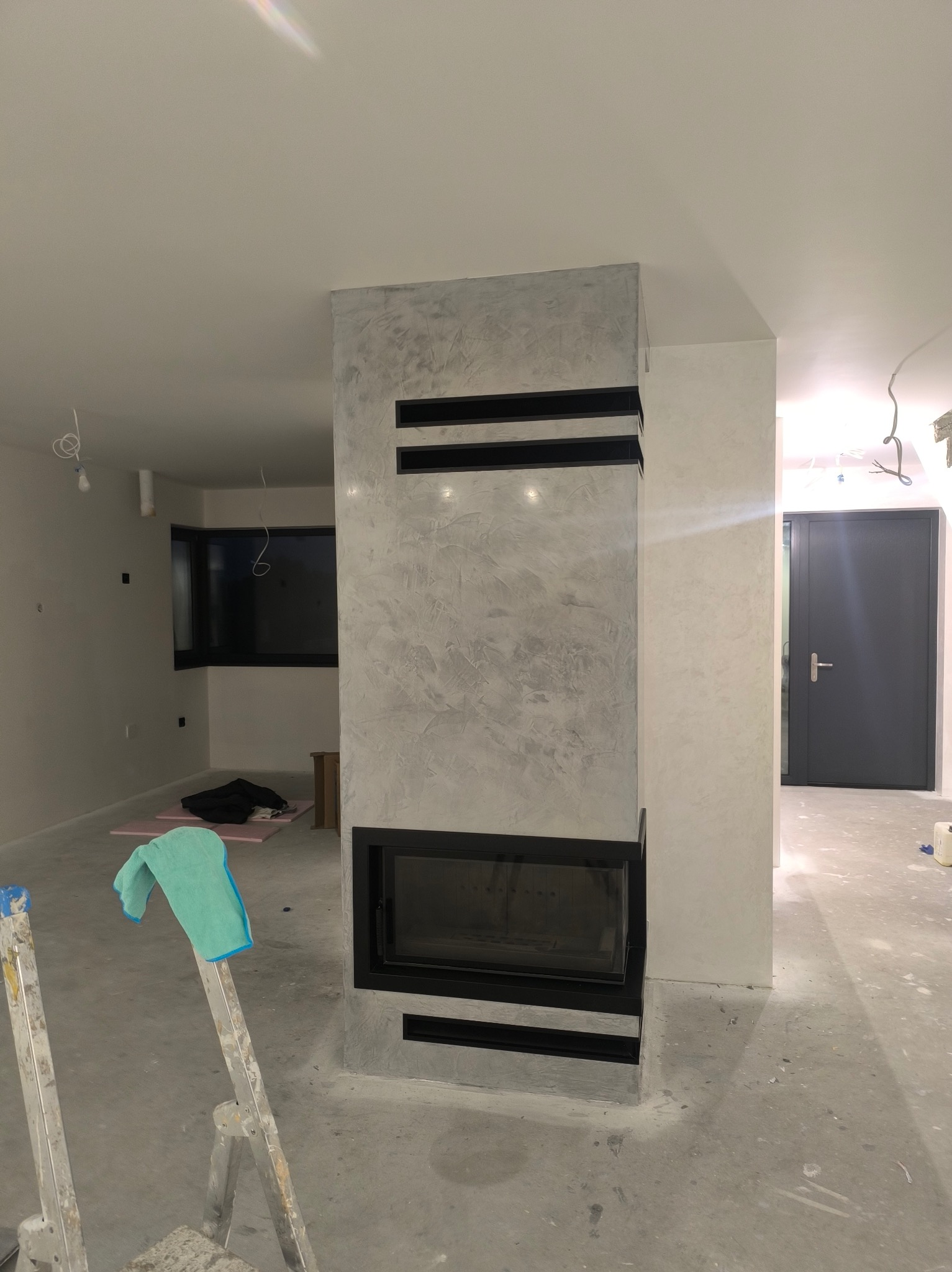 Wykończony kominek z dekoracyjnym tynkiem w szarościach, czarne elementy ozdobne. W tle widoczne okno i drzwi, pomieszczenie w trakcie wykańczania.