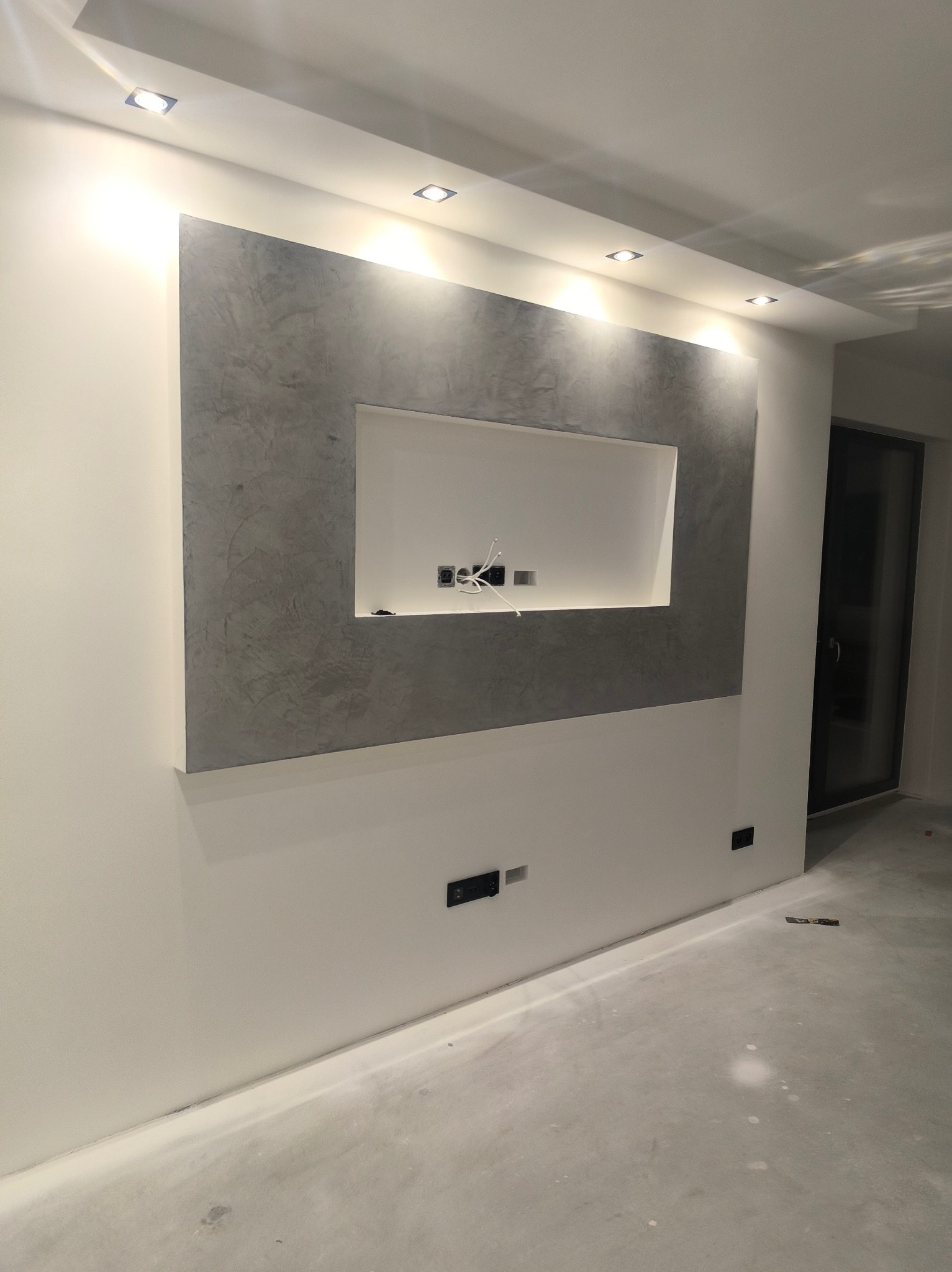 Ściana z dekoracyjnym tynkiem w odcieniach szarości, z wnęką na telewizor i punktami świetlnymi w suficie. Widoczne gniazdka elektryczne i surowa podłoga.