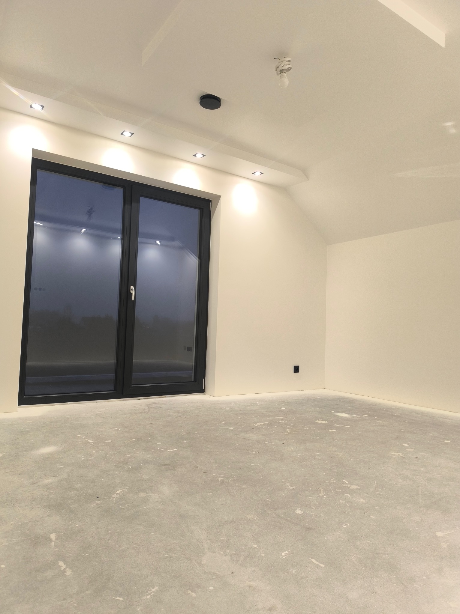 Wnętrze po remoncie w Silnicy: białe ściany, szara podłoga, czarne okno. Widoczne punktowe oświetlenie sufitowe i gładkie powierzchnie. Minimalistyczny design.