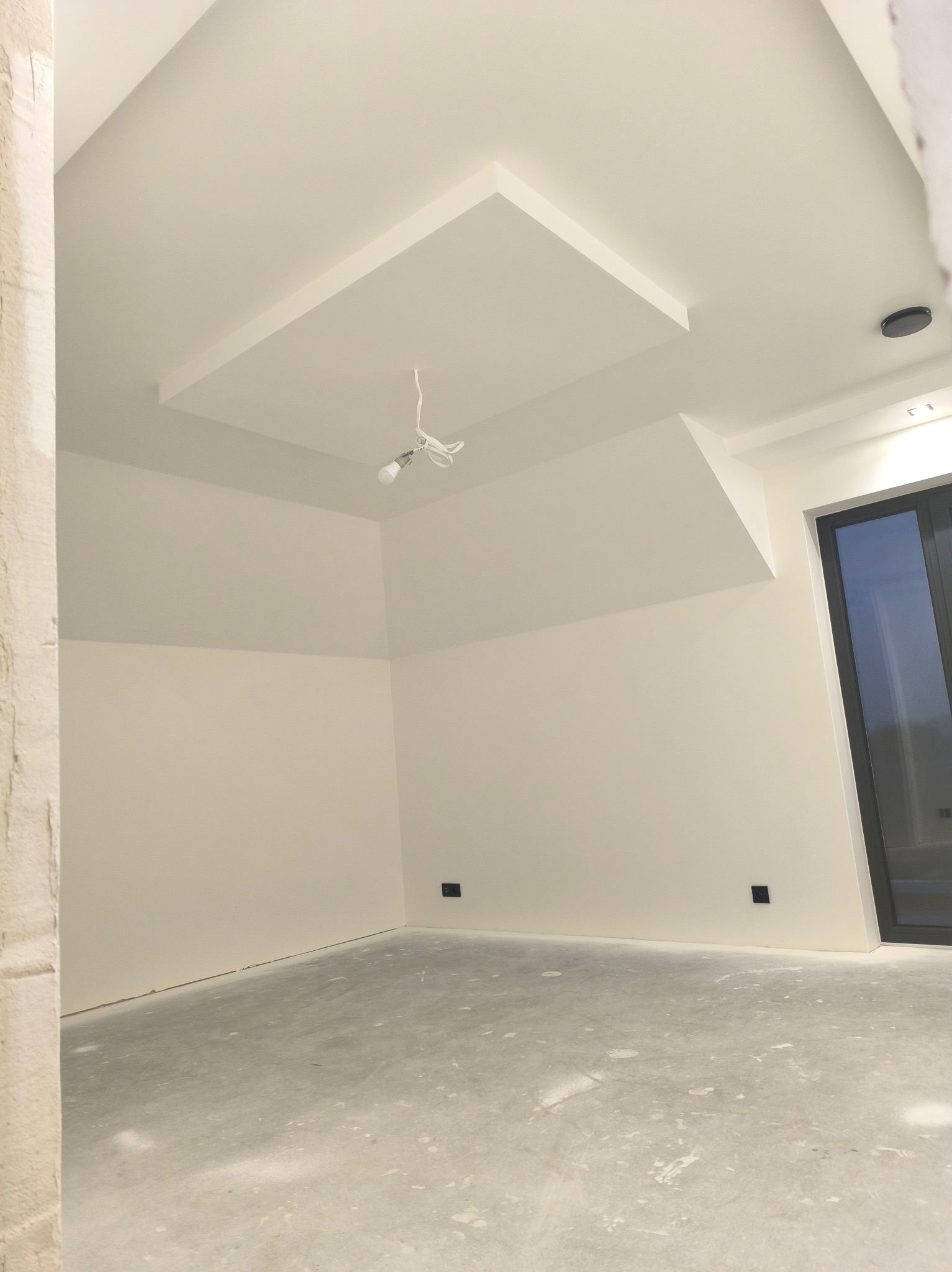 Jasne, świeżo pomalowane wnętrze z surową podłogą i nowoczesnym oświetleniem sufitowym. Widoczne gniazdka elektryczne i duże okno przesuwne.