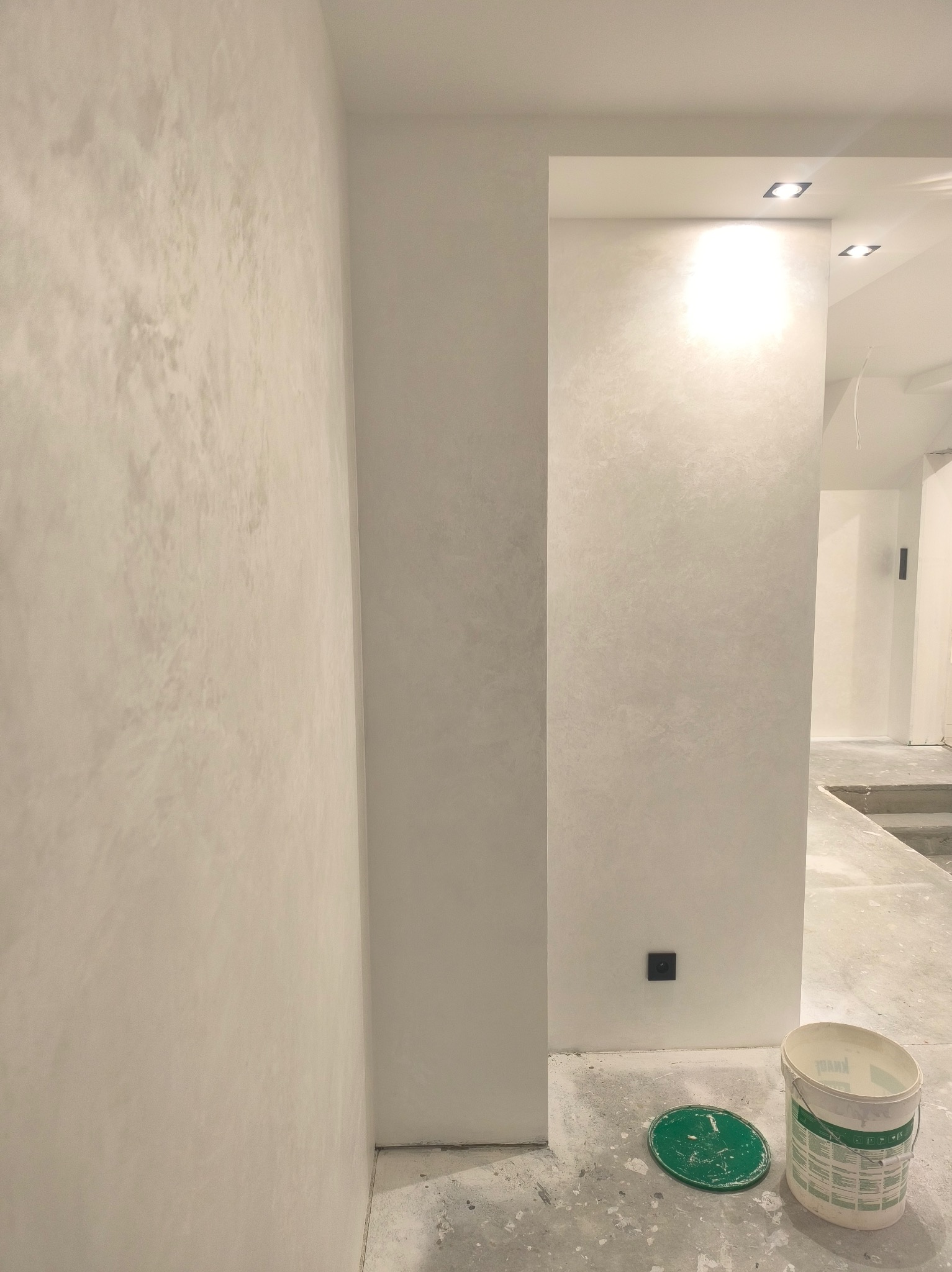Ściana z dekoracyjnym tynkiem w odcieniach szarości, widoczny fragment podłogi z wiadrem po farbie i gniazdko elektryczne. W tle przejście do innego pomieszczenia.