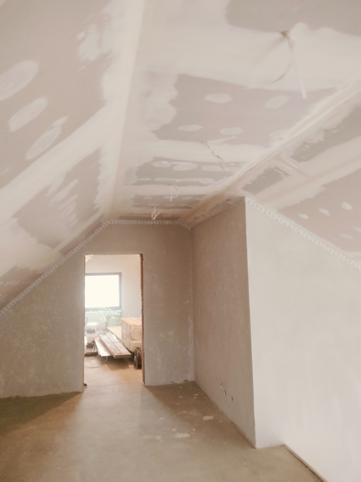 Poddasze w trakcie wykańczania: ściany i sufit pokryte gładzią gipsową, widoczne kable, listwy dekoracyjne pod sufitem, otwarte przejście do kolejnego pomieszczenia z ułożonymi deskami.