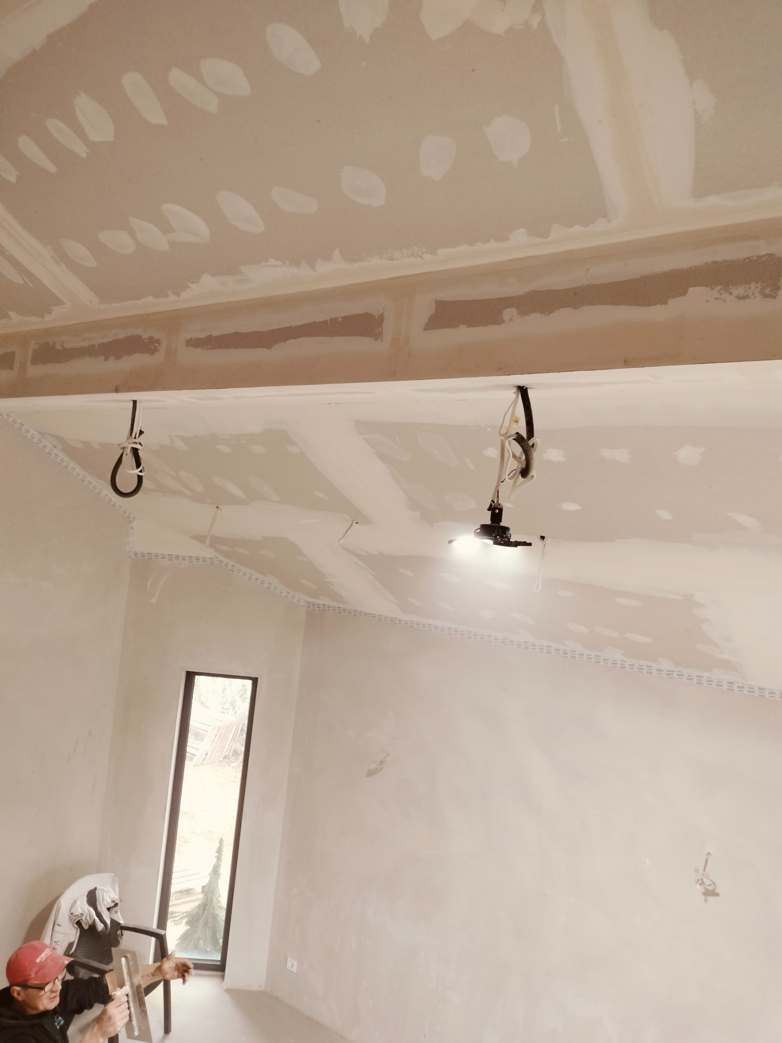 Wnętrze w trakcie prac wykończeniowych; widoczny pracownik z kielnią szpachlową, szpachlowana ściana i sufit z widocznymi łączeniami płyt gipsowo-kartonowych oraz lampa budowlana na statywie.
